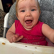 Aliyah is registered to the contest to win money with this photo: baby, child, high_chair, pink_shirt, happy, smiling, food, crumbs, face, mouth_open, indoor, person, seat, feeding, cute, expression, hair, table, furniture, living_room