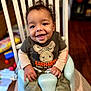 toddler, baby_seat, smiling, child, happy, clothing, camouflage, carhartt, indoor, furniture, wood_floor, toy, hair, face, person, sitting, cute, young_child, home, playful