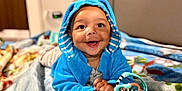 Noah is registered to the contest to win money with this photo: baby, child, blue_hoodie, smiling, toy, blanket, indoor, portrait, happy, cute, infant, clothing, playful, face, hands, toddler, bedroom, cozy, colorful, lying_down