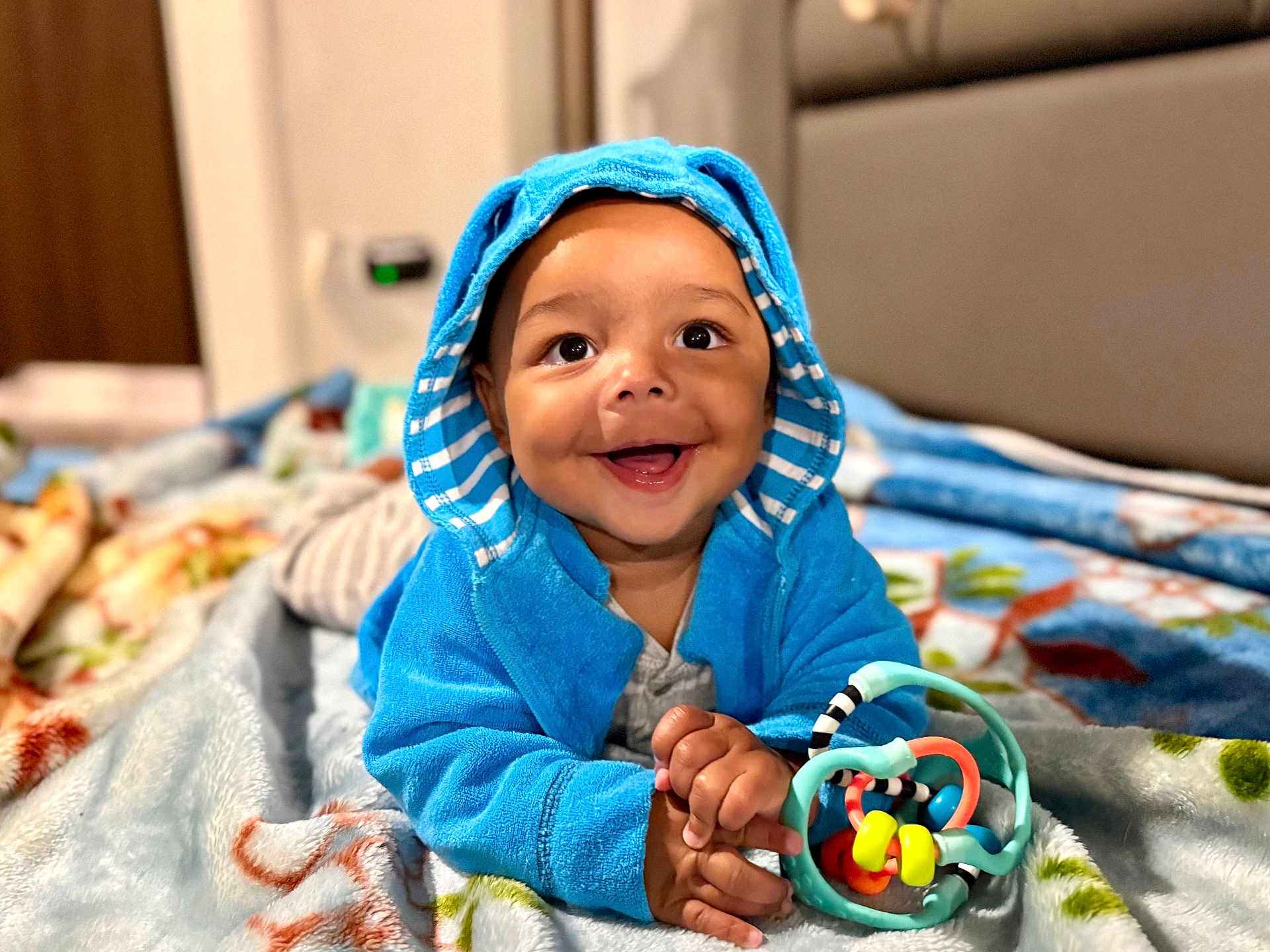 Noah is registered to the contest to win money with this photo: baby, child, blue_hoodie, smiling, toy, blanket, indoor, portrait, happy, cute, infant, clothing, playful, face, hands, toddler, bedroom, cozy, colorful, lying_down
