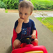 Matthew is registered to the contest to win money with this photo: toddler, child, toy_car, red, blue_shirt, stars, outdoor, greenery, concrete, hand, fingers_in_mouth, shorts, steering_wheel, young_child, summer, playtime, daytime, casual_clothing, expression, person