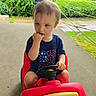 toddler, child, toy_car, red, blue_shirt, stars, outdoor, greenery, concrete, hand, fingers_in_mouth, shorts, steering_wheel, young_child, summer, playtime, daytime, casual_clothing, expression, person