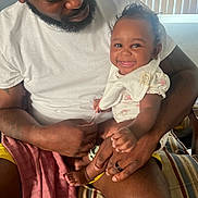Olivia joined the competition — help win amazing prizes! baby, adult, smile, indoor, lap, clothing, beard, hat, curly_hair, window_blinds, person, happy, holding, skin, furniture, bib, shorts, tshirt, seated, home
