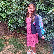 Brooklin is registered to the contest to win money with this photo: child, girl, smiling, backpack, dress, cardigan, shoes, grass, bushes, outdoor, plants, greenery, happy, portrait, standing, young, casual_clothing, daylight, nature, cute