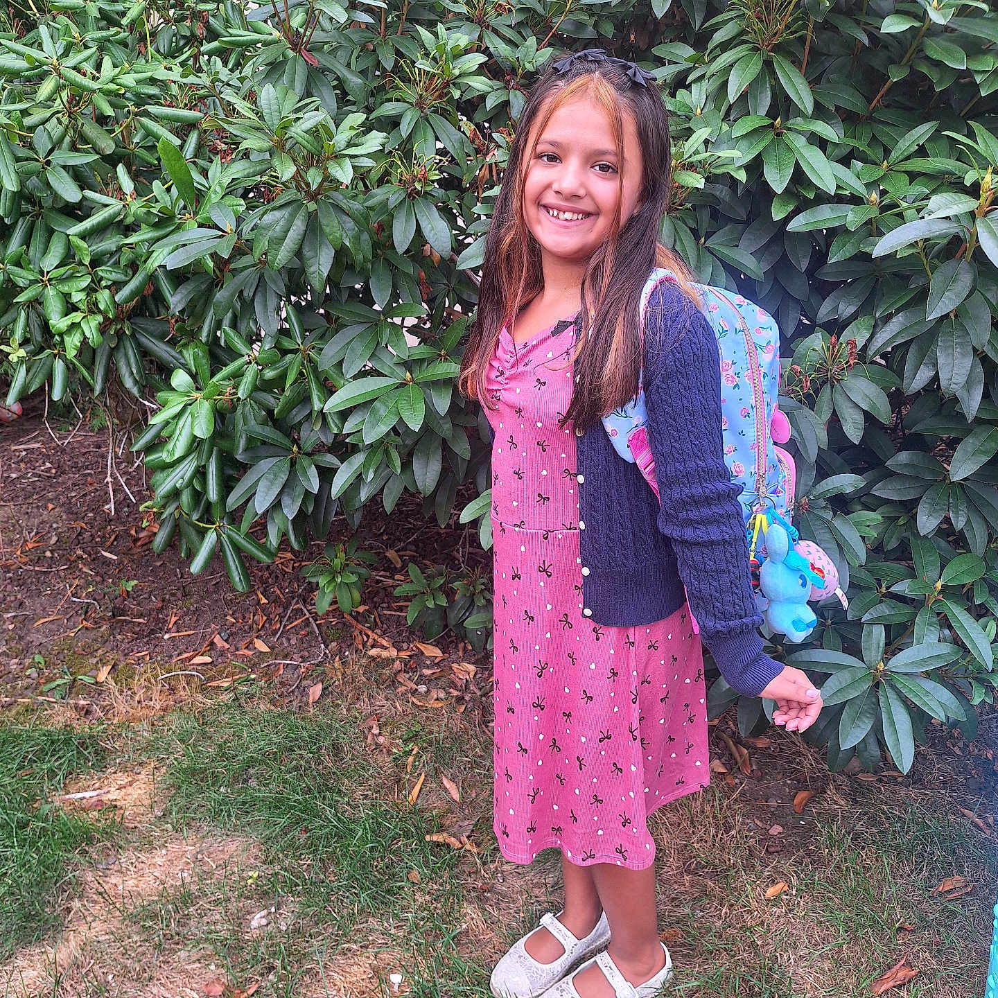 Brooklin is registered to the contest to win money with this photo: backpack, bushes, cardigan, casual_clothing, child, cute, daylight, dress, girl, grass, greenery, happy, nature, outdoor, plants, portrait, shoes, smiling, standing, young