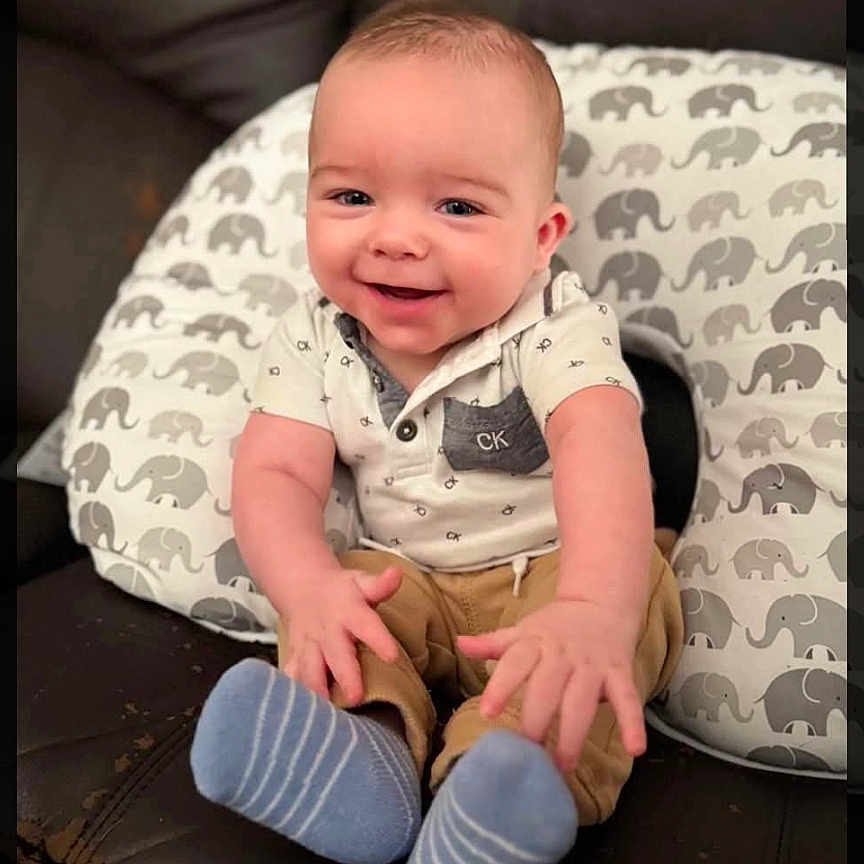 Declan joined the competition — help win amazing prizes! baby, smiling, infant, sitting, couch, pillow, elephant_pattern, blue_socks, white_shirt, beige_pants, happy, child, indoor, portrait, cute, young, person, comfortable, leather, home