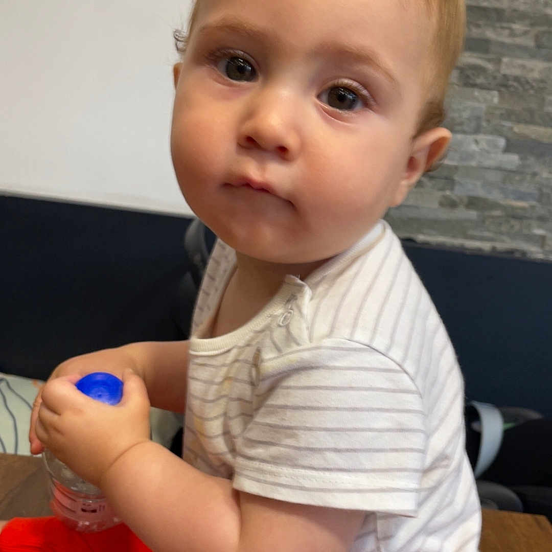 Brent a rejoint le concours — aidez-le/la à gagner de superbes lots ! baby, casual_wear, child, clothing, couch, curious, expression, face, hand, indoor, innocent, person, red_pants, sitting, stone_wall, striped_shirt, toddler, water_bottle, wooden_table, young_child
