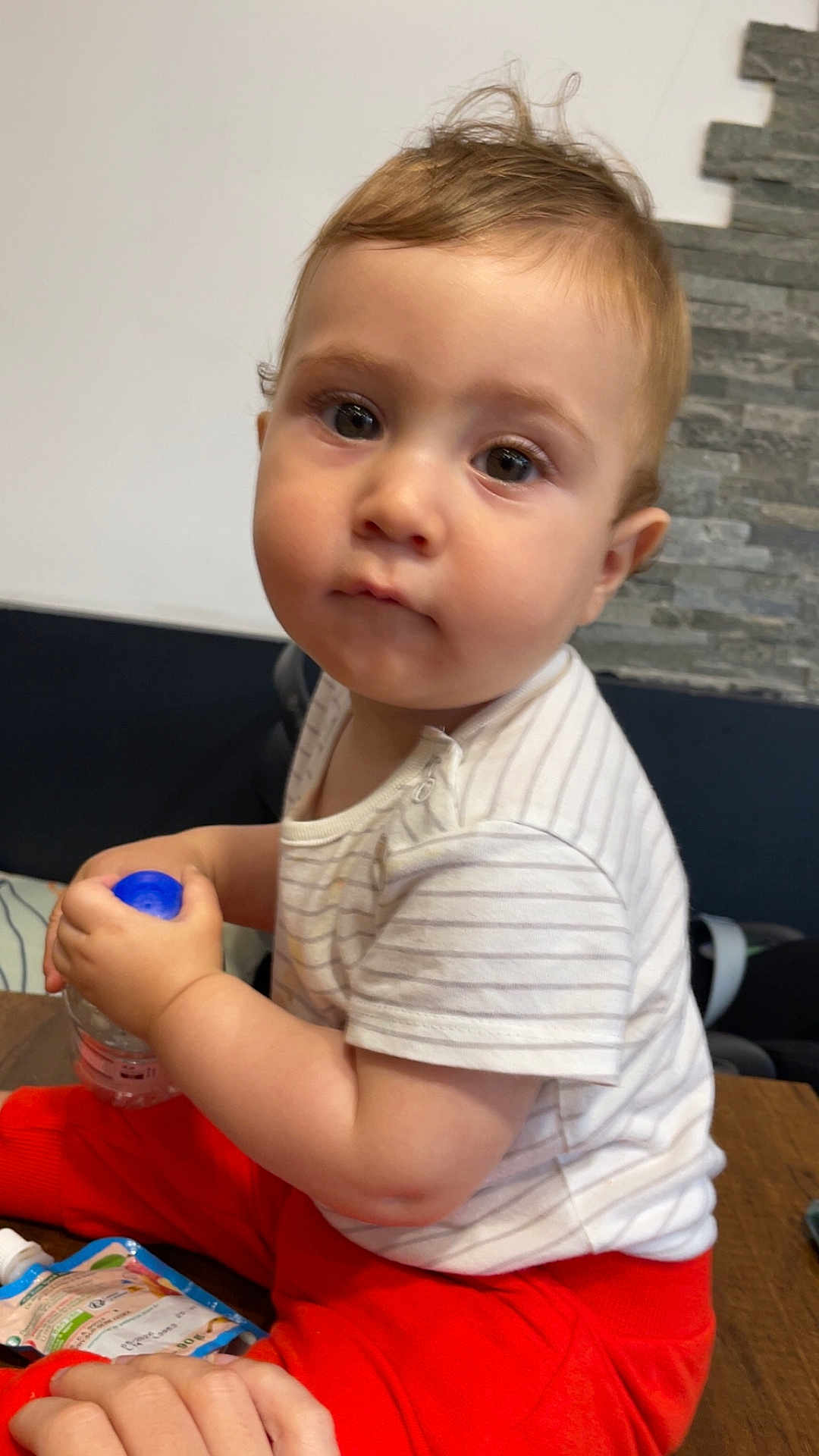 Brent a rejoint le concours — aidez-le/la à gagner de superbes lots ! baby, toddler, child, water_bottle, striped_shirt, red_pants, indoor, wooden_table, curious, face, sitting, person, hand, clothing, couch, stone_wall, innocent, expression, young_child, casual_wear