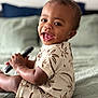 Zyaire is registered to the contest to win money with this photo: toddler, child, smiling, happy, sitting, bed, blanket, clothing, palm_tree_pattern, indoor, person, cute, portrait, baby, skin, hand, face, eyes, mouth, hair