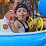 child, adult, amusement_ride, blue, yellow, steering_wheel, cap, tongue_out, glasses, woman, lights, carousel, fun, playful, outdoor, person, face, hair, clothing, seat