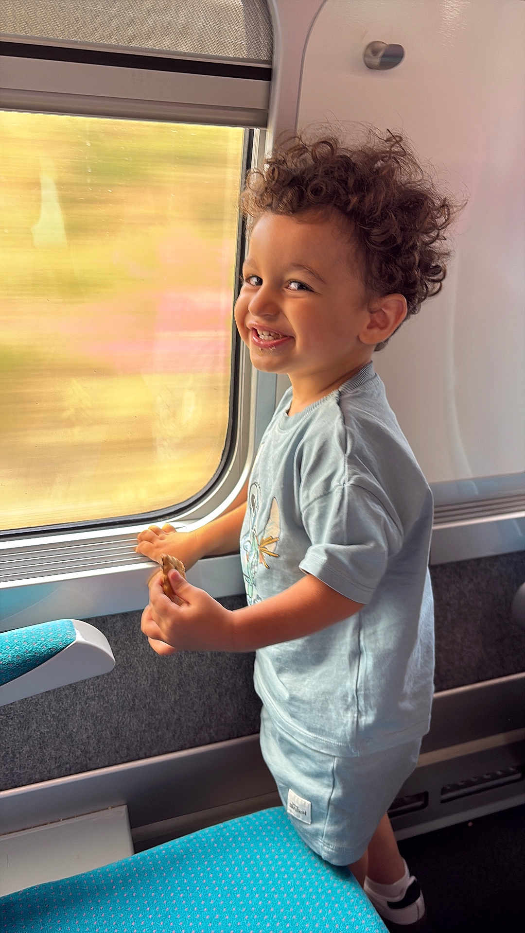 Lenzo a rejoint le concours — aidez-le/la à gagner de superbes lots ! child, toddler, curly_hair, smiling, train, window, snack, seat, clothing, shorts, tshirt, indoor, travel, motion_blur, happy, person, footwear, hand, side_view, young