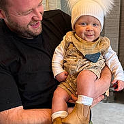Finley is registered to the contest to win money with this photo: man, baby, child, smile, hat, boots, bib, indoor, holding, happy, beard, black_shirt, light_skin, short_hair, casual_clothing, portrait, warm_lighting, cozy, infant, parent