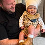 man, baby, child, smile, hat, boots, bib, indoor, holding, happy, beard, black_shirt, light_skin, short_hair, casual_clothing, portrait, warm_lighting, cozy, infant, parent