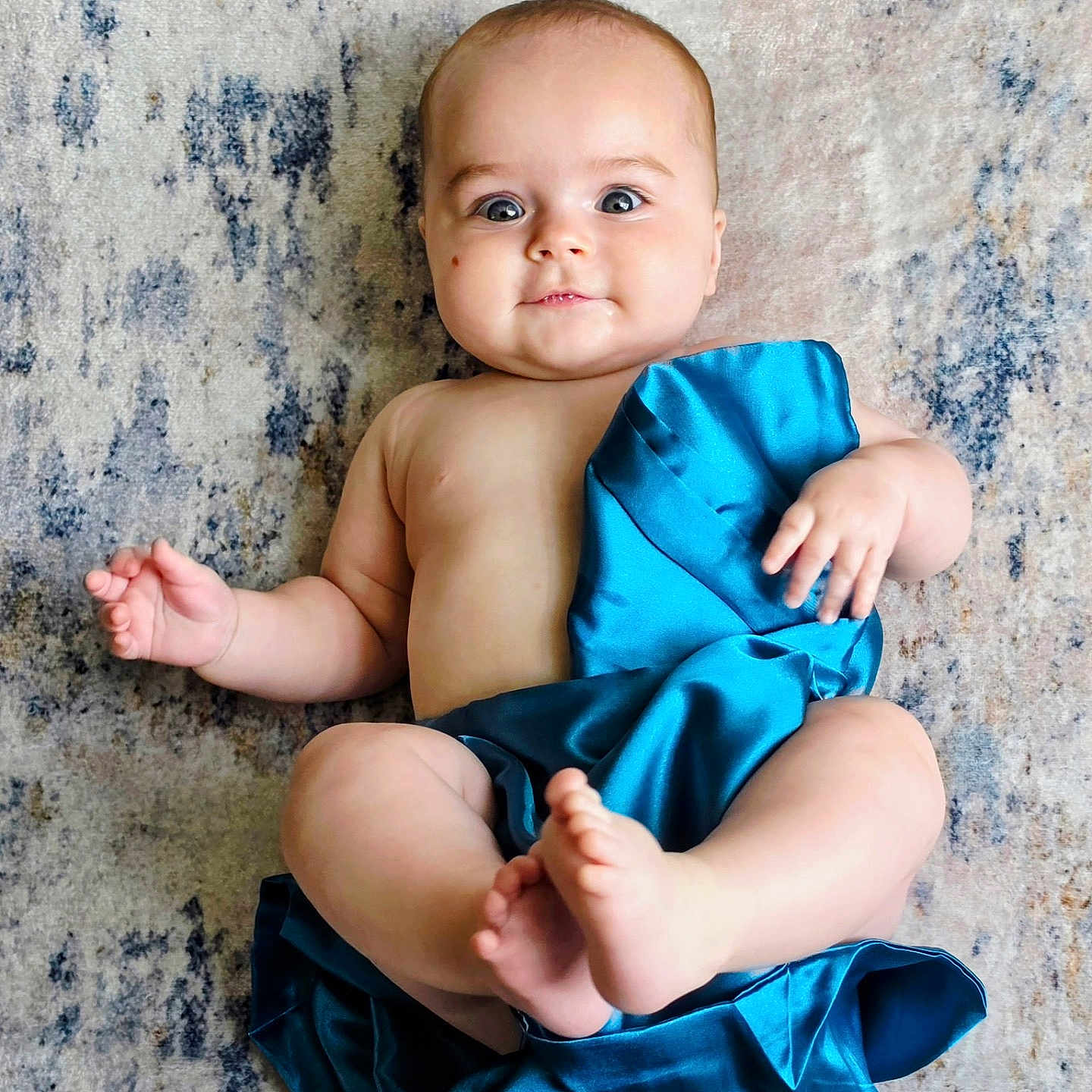 Levi is registered to the contest to win money with this photo: baby, infant, child, blue_cloth, bare_skin, lying_down, rug, floor, curious_expression, wide_eyes, chubby_cheeks, soft_texture, indoor, cute, portrait, human, small_hands, small_feet, skin, relaxed