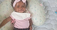Bless'In is registered to the contest to win money with this photo: baby, child, infant, smiling, portrait, pink_bow, headband, curly_hair, dark_skin, baby_clothing, shirt, shorts, socks, pillow, floral_pillow, bed, quilt, stuffed_toy, toy, indoor