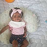 baby, child, infant, smiling, portrait, pink_bow, headband, curly_hair, dark_skin, baby_clothing, shirt, shorts, socks, pillow, floral_pillow, bed, quilt, stuffed_toy, toy, indoor