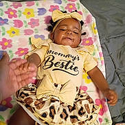 Bless'In is registered to the contest to win money with this photo: baby, infant, child, portrait, lying_down, smiling, headband, bow, dress, leopard_print, floral_blanket, bed, bedroom, adult_hand, fashion, cute, face, person, tiktok_watermark, indoor