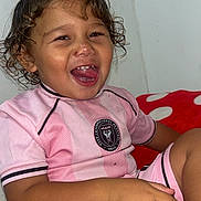 Pablo Ruiz participe au concours pour gagner de l'argent avec cette photo : blanket, casual, child, curly_hair, cute, face, hand, happy, indoor, legs, person, pink_clothing, red, sitting, smiling, soccer_jersey, toddler, wall, white, young_child