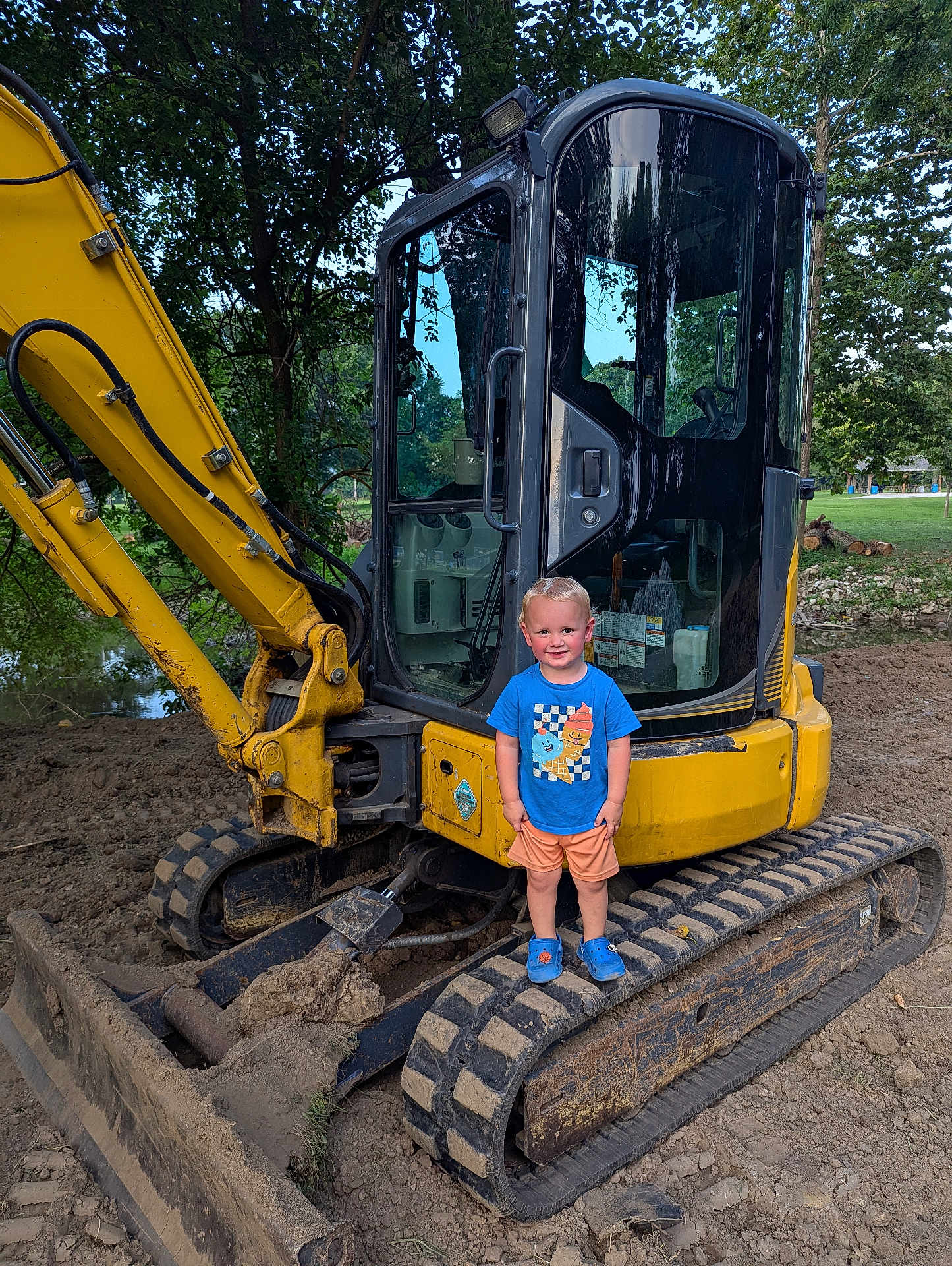 Rhett joined the competition — help win amazing prizes! child, toddler, excavator, construction, mud, yellow, blue_shirt, orange_shorts, blue_shoes, outdoor, dirt, trees, smiling, heavy_machinery, playful, equipment, vehicle, person, sunlight, nature