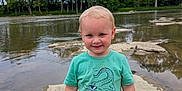 Rhett is registered to the contest to win money with this photo: child, toddler, boy, smiling, standing, shirt, crocodile_graphic, shorts, outdoor, river, rocks, water, trees, nature, daylight, sky, casual_clothing, happy, portrait, playful