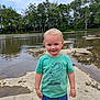 child, toddler, boy, smiling, standing, shirt, crocodile_graphic, shorts, outdoor, river, rocks, water, trees, nature, daylight, sky, casual_clothing, happy, portrait, playful