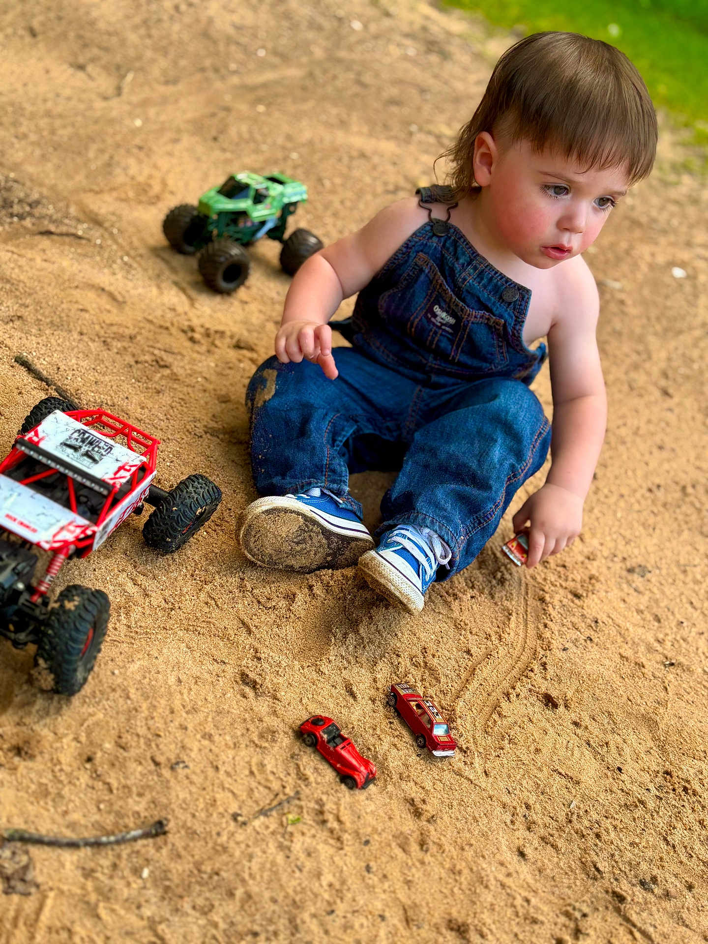 Daxton joined the competition — help win amazing prizes! toddler, child, overalls, denim, sneakers, toy_cars, monster_truck, sand, outdoor, play, grass, closeup, casual, childhood, young_child, person, cute, focused, sitting, daylight
