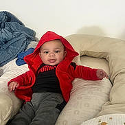 Oliver is registered to the contest to win money with this photo: baby, blanket, child, clothing, cozy, cushion, cute, face, furniture, gray_pants, happy, home, indoor, infant, person, pillow, red_hoodie, relaxed, smile, tongue_out