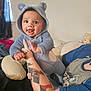 arm, baby, blue_onesie, child, childrens_clothing, cozy, cute, face, furniture, hand, happy, hood, indoor, infant, person, pillow, room, smile, tattoo, teddy_bear