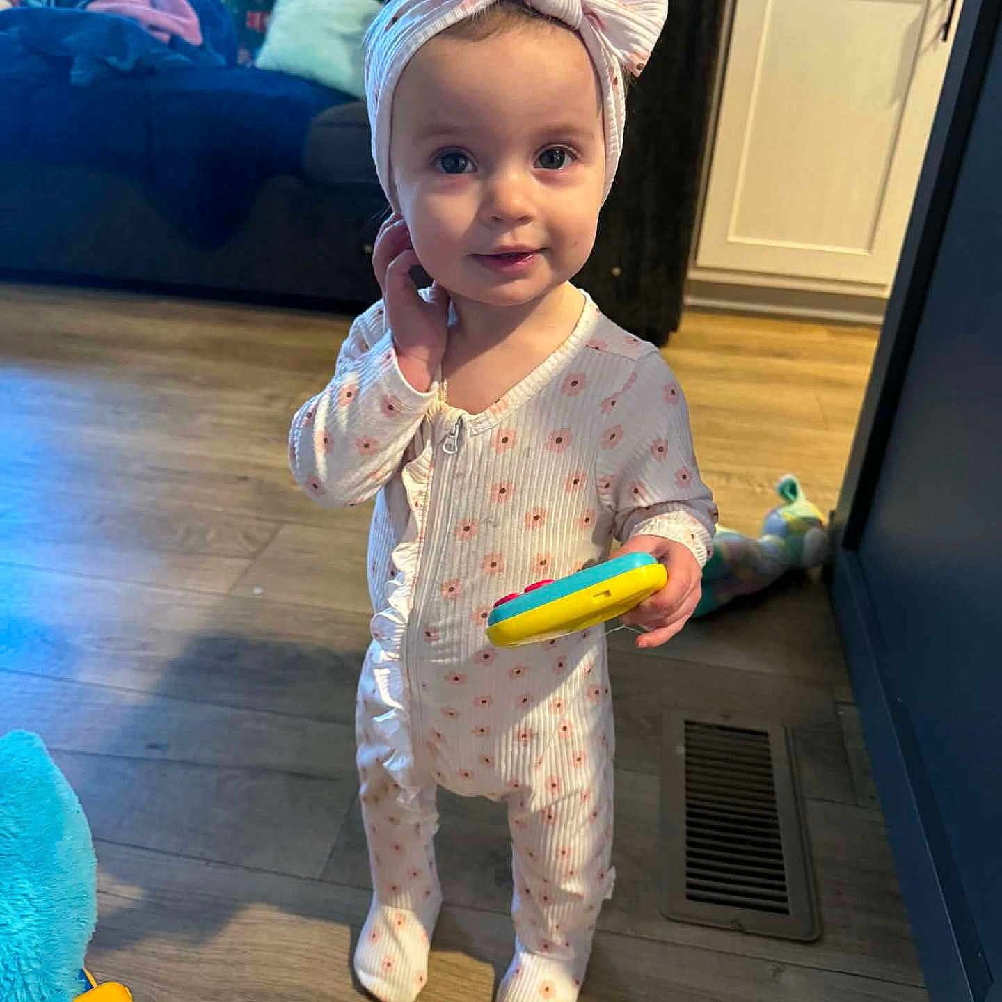 Jocelynne joined the competition — help win amazing prizes! baby, child, toddler, standing, smiling, portrait, headband, bow, pajamas, floral_pattern, toy, colorful_toy, indoors, living_room, sofa, wooden_floor, laundry, cabinet, home, family_photo