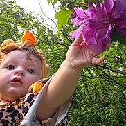 Lavanna joined the competition — help win amazing prizes! toddler, child, flower, purple_flower, orange_headband, nature, greenery, outdoor, sunlight, trees, leaves, curious, reaching_hand, baby, dress, patterned_clothing, person, portrait, summer, garden