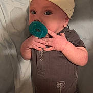 Augustus is registered to the contest to win money with this photo: baby, infant, pacifier, hat, onesie, guitar_print, hands, face, eyes, lying_down, bed, blanket, clothing, cute, child, newborn, skin, expression, indoors, soft_surface