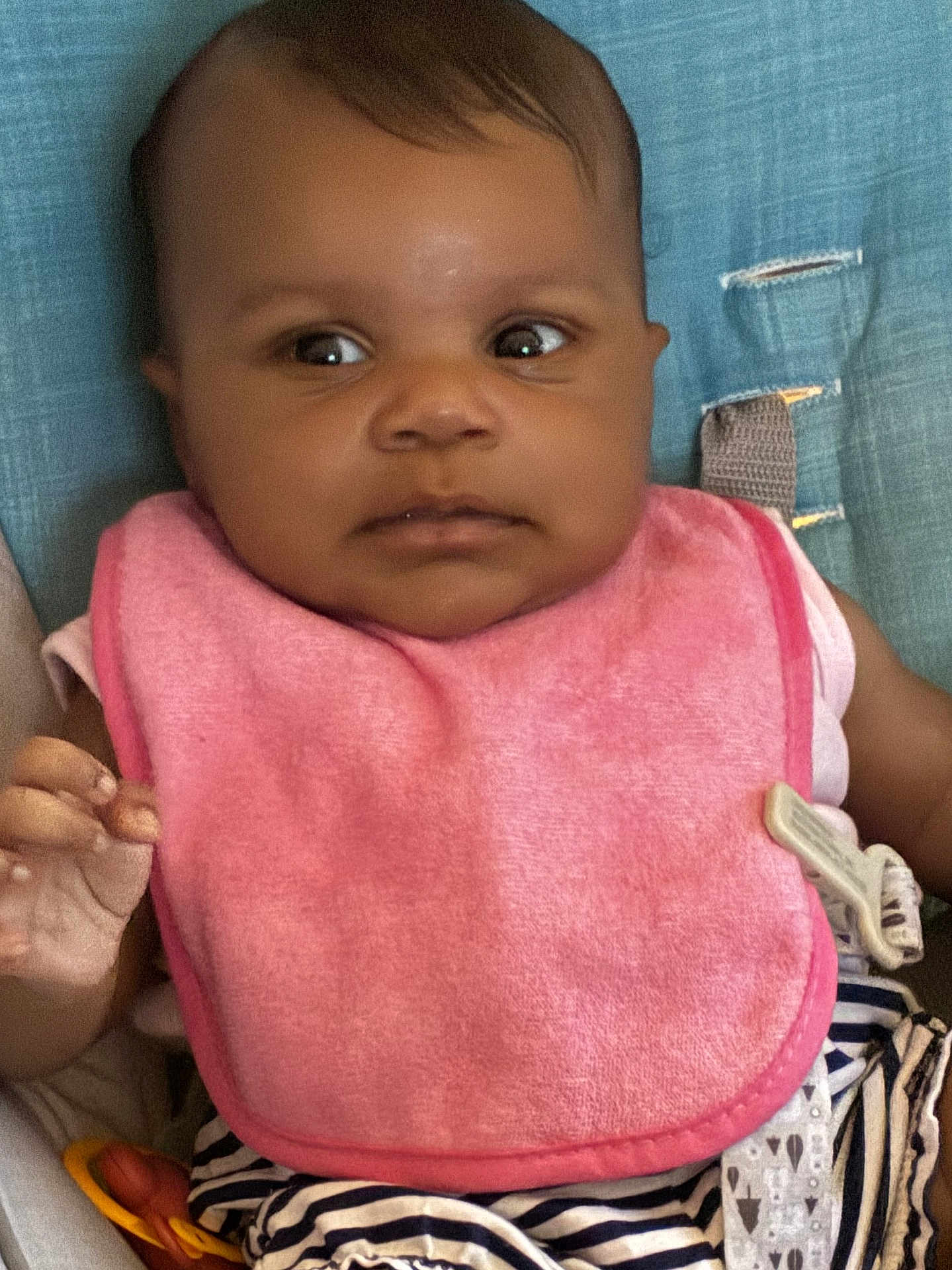 Keziah joined the competition — help win amazing prizes! baby, infant, pink_bib, striped_pants, curious, face, hand, blue_background, clothing, sitting, toy, indoors, child, person, closeup, young, cute, expression, portrait, seat
