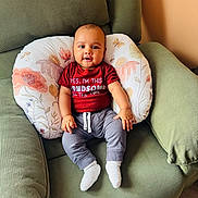 Noah joined the competition — help win amazing prizes! baby, child, infant, red_shirt, gray_pants, white_socks, green_chair, floral_pillow, indoor, smiling, seated, cute, comfortable, portrait, person, home, wooden_floor, handsome, young, casual_clothing