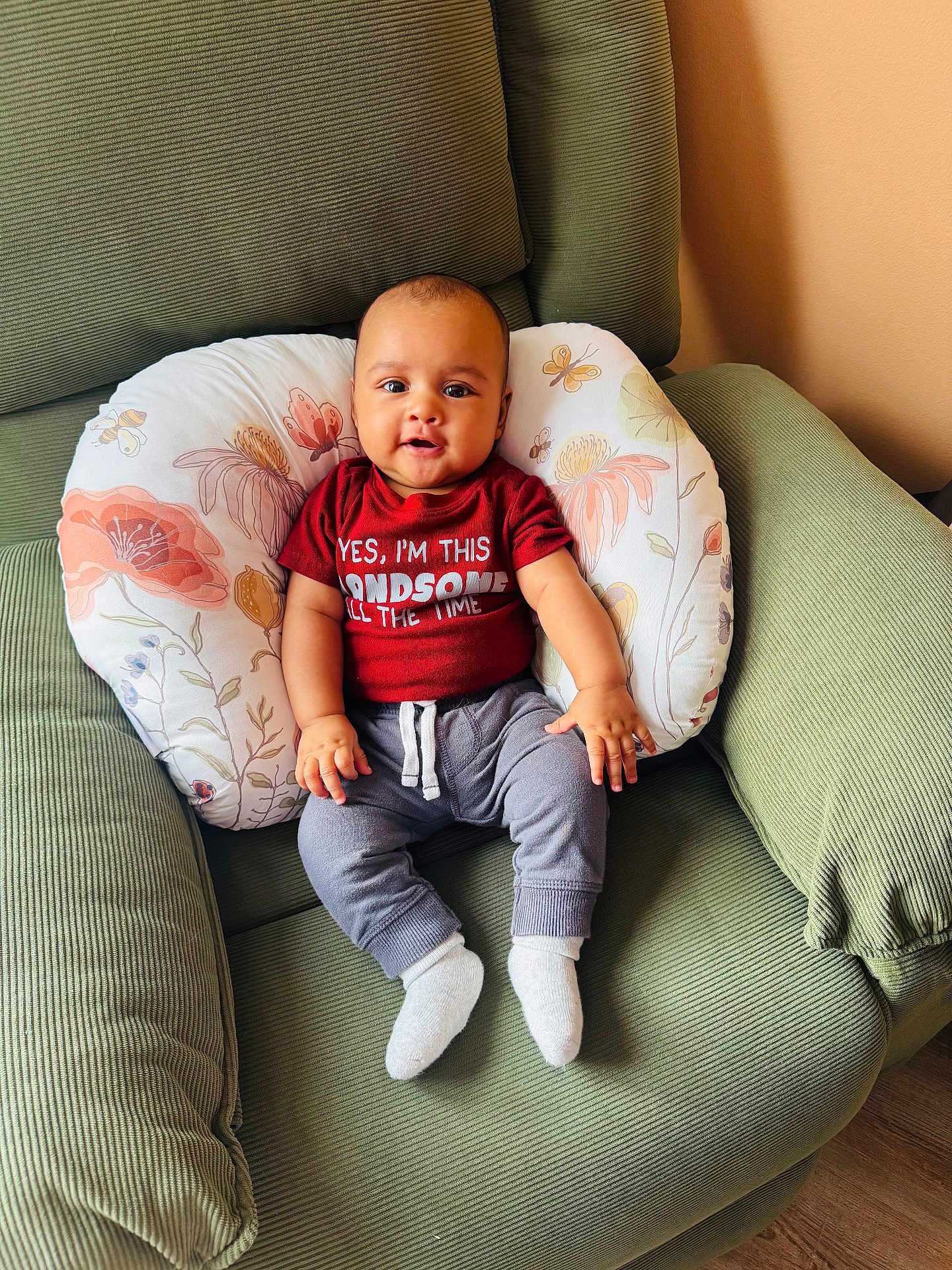 Noah joined the competition — help win amazing prizes! baby, child, infant, red_shirt, gray_pants, white_socks, green_chair, floral_pillow, indoor, smiling, seated, cute, comfortable, portrait, person, home, wooden_floor, handsome, young, casual_clothing