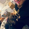 man, cat, smile, indoor, cozy, blanket, pillow, tabby_cat, casual, seated, home, furniture, cardboard_box, facial_hair, black_clothing, pet, relaxed, window, curtains, friendly