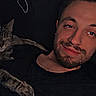 Jade is registered to the contest to win money with this photo: cat, man, indoor, relaxed, smile, sleepy, pet, companion, dark_background, casual_clothing, beard, feline, resting, couch, friendship, comfort, domestic, portrait, evening, closeup