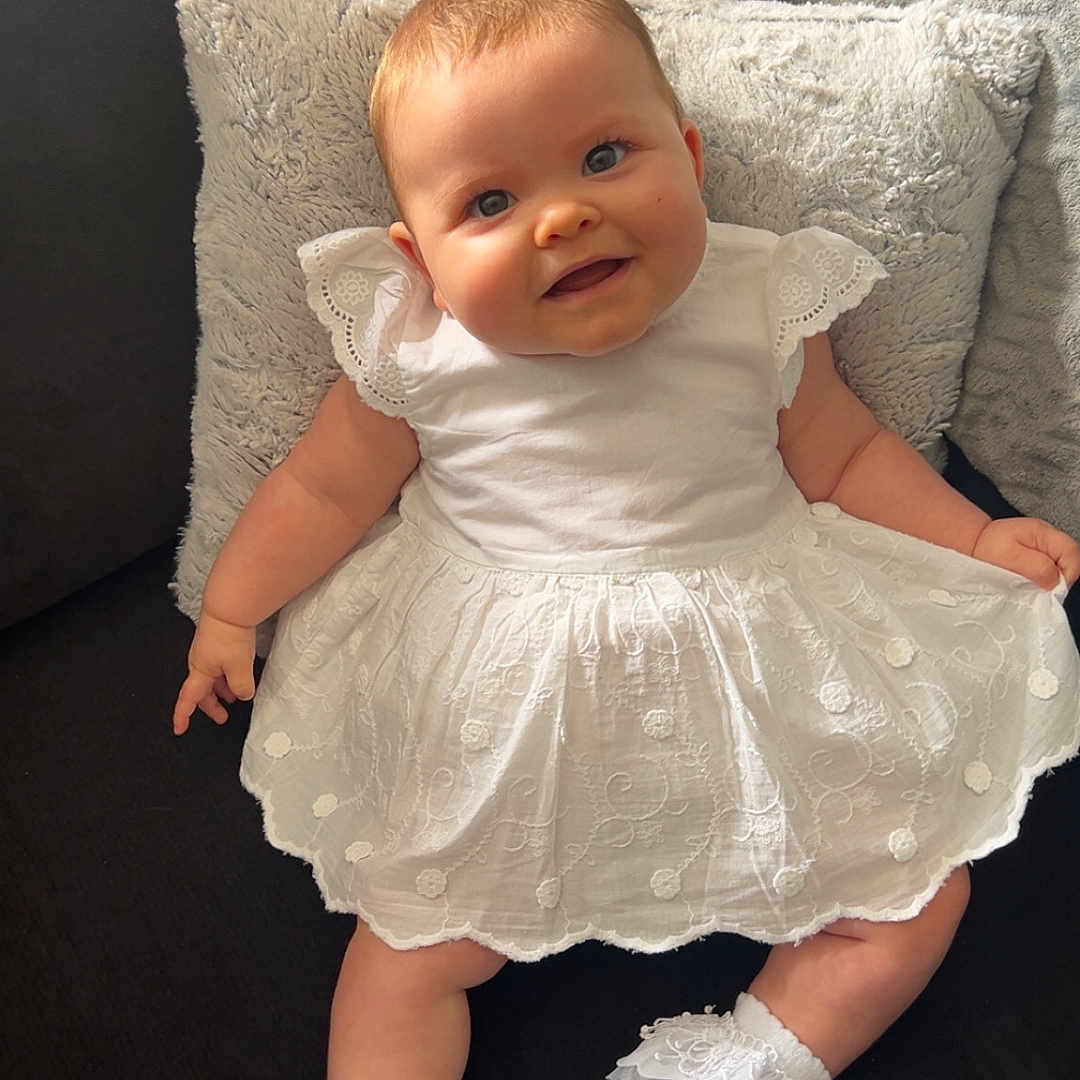 Sophia joined the competition — help win amazing prizes! baby, child, couch, cozy, cute, fabric, happy, indoor, infant, lace, person, pillow, plush, portrait, sitting, smile, socks, soft_texture, white_dress, young