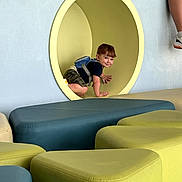 Gunner is registered to the contest to win money with this photo: toddler, child, playground, indoor, crawling, yellow_tunnel, backpack, blue, green, cushions, seated, legs, smiling, play_area, fun, casual_clothing, camouflage_shorts, socks, shoes, wall