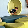 toddler, child, playground, indoor, crawling, yellow_tunnel, backpack, blue, green, cushions, seated, legs, smiling, play_area, fun, casual_clothing, camouflage_shorts, socks, shoes, wall