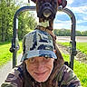 brown_dog, camouflage, collar, dog, greenery, happy, hat, hoodie, man, nature, outdoor, pet, portrait, road, selfie, shoulders, sky, smile, sunny, trees