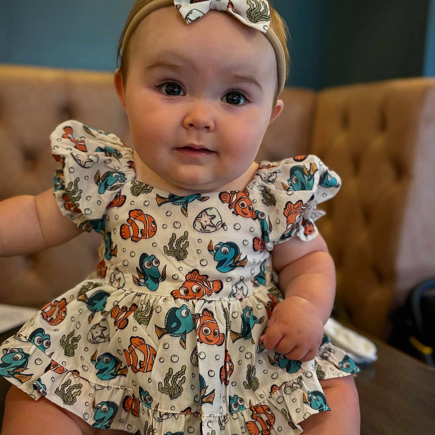 Reeva joined the competition — help win amazing prizes! baby, bow, bracelet, child, chubby_cheeks, clothing, couch, cute, dress, fish_pattern, furniture, happy, headband, indoor, infant, person, portrait, sitting, smiling, table