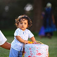Maghathi joined the competition — help win amazing prizes! toddler, child, curly_hair, toy, stool, flamingo_pattern, grass, outdoor, person, hand, blue_shorts, white_shirt, basket, nature, daylight, portrait, playing, support, kneeling_adult, background_blur