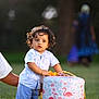 toddler, child, curly_hair, toy, stool, flamingo_pattern, grass, outdoor, person, hand, blue_shorts, white_shirt, basket, nature, daylight, portrait, playing, support, kneeling_adult, background_blur