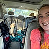 woman, smile, selfie, car_interior, puppy, golden_retriever, blanket, seat, headrest, hat, sunlight, collar, travel, young_adult, happy, casual_clothing, window, daytime, leather_seat, accessory