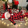 baby, child, christmas_tree, christmas_lights, santa_hat, plaid_onesie, gift_boxes, wooden_floor, blanket, holiday_decor, ornament, festive, indoors, holiday_season, cute, smiling, person, celebration, warm_lighting, decorations