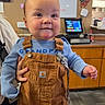baby, child, overalls, blue_shirt, smile, indoor, person, table, pacifier, hand, face, curly_hair, clothing, background, counter, computer_screen, decor, wall_art, adult_hand, floor