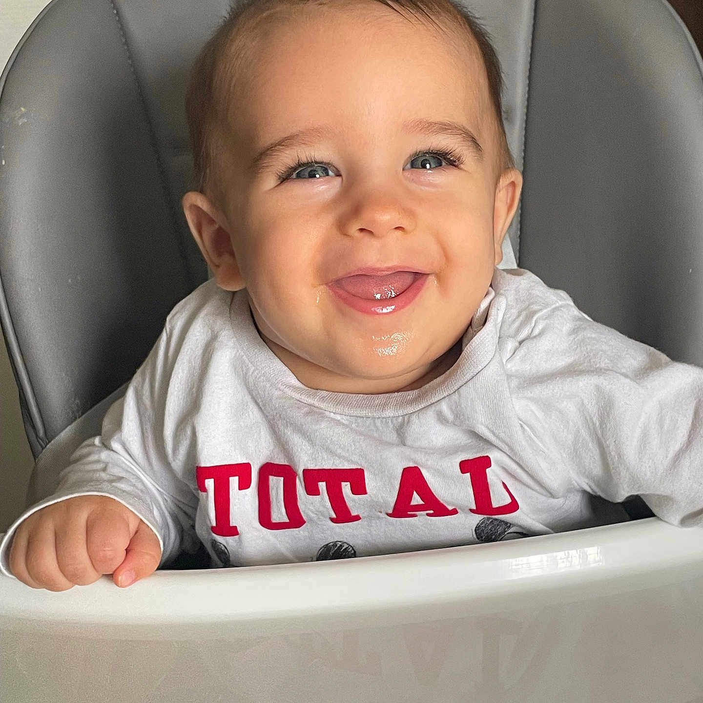 Ezïo participe au concours pour gagner de l'argent avec cette photo : baby, child, clothing, cute, eyes, face, happy, high_chair, indoor, infant, joyful, person, portrait, red_letters, seat, skin, smile, tongue, white_shirt, young_child