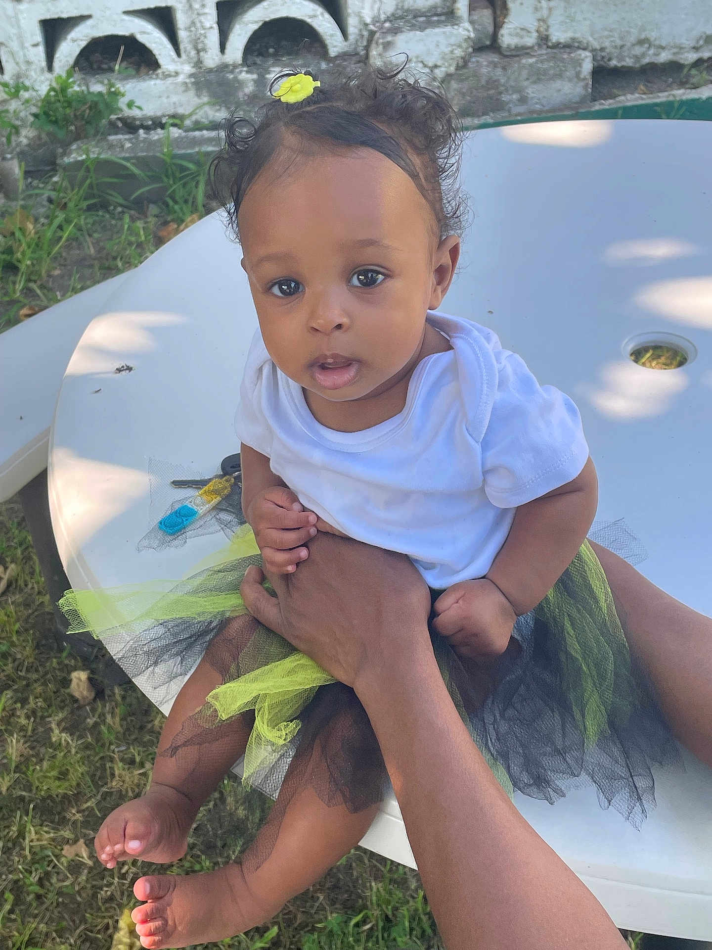 Saiaura is registered to the contest to win money with this photo: baby, child, outdoor, table, white_shirt, tutu, flower_hair_clip, grass, hands, person, curious, skin, feet, legs, holding, sunlight, shadow, nature, face, portrait