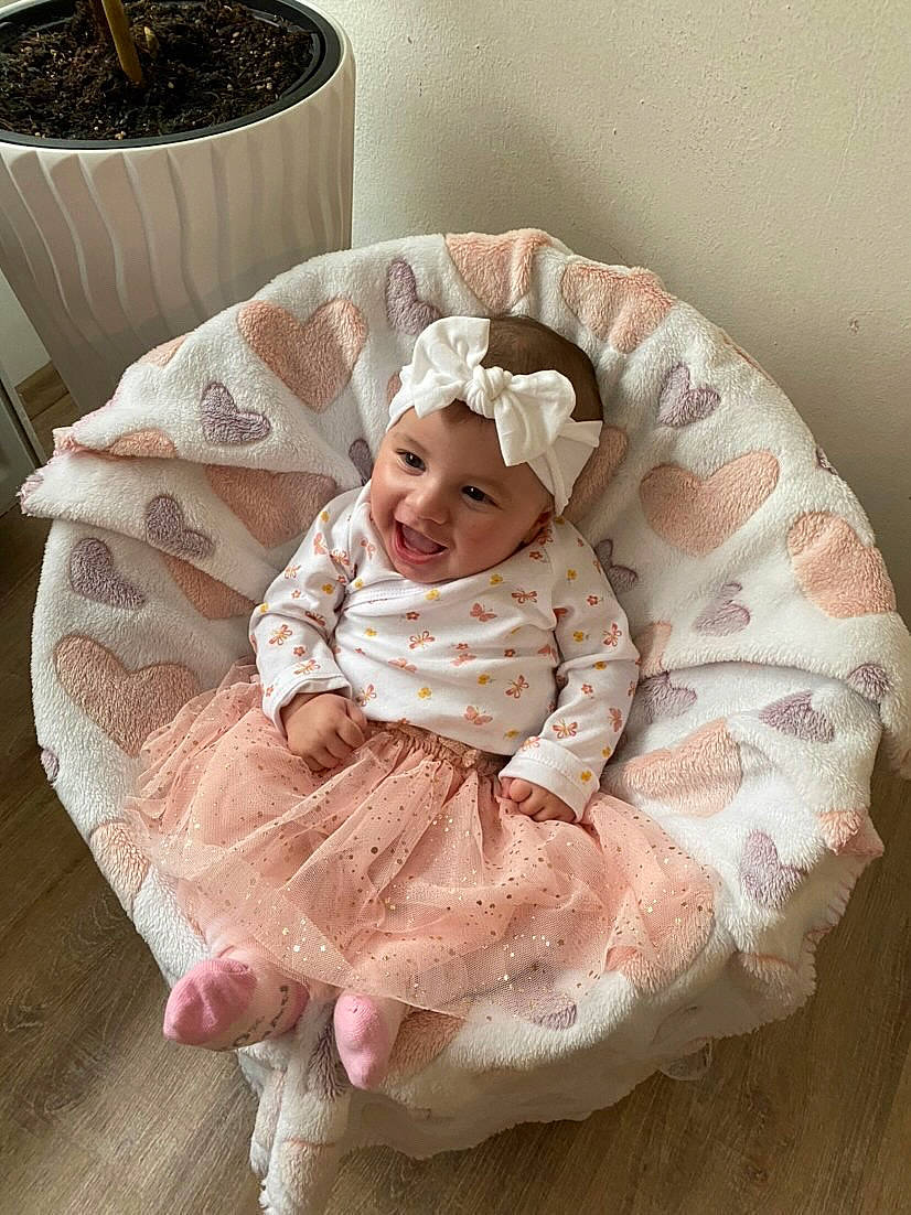 Talia participe au concours pour gagner de l'argent avec cette photo : baby, baby_products, baby_toddler_clothing, child, clothing, comfort, eye, face, flooring, hair_accessory, head, headgear, headwear, joy, linens, peach, person, pink, sitting, sleeve