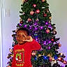 child, toddler, christmas_tree, christmas, holiday, ornaments, lights, star, grinch, pajamas, red_slippers, smiling, salute, standing, indoor, home, festive, portrait, decorations, tree_skirt